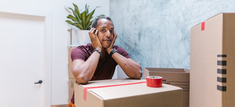 A man resting on a cardboard box that has a packing tape on it, which are the necessary packing supplies to prepare a flatscreen TV for your move.