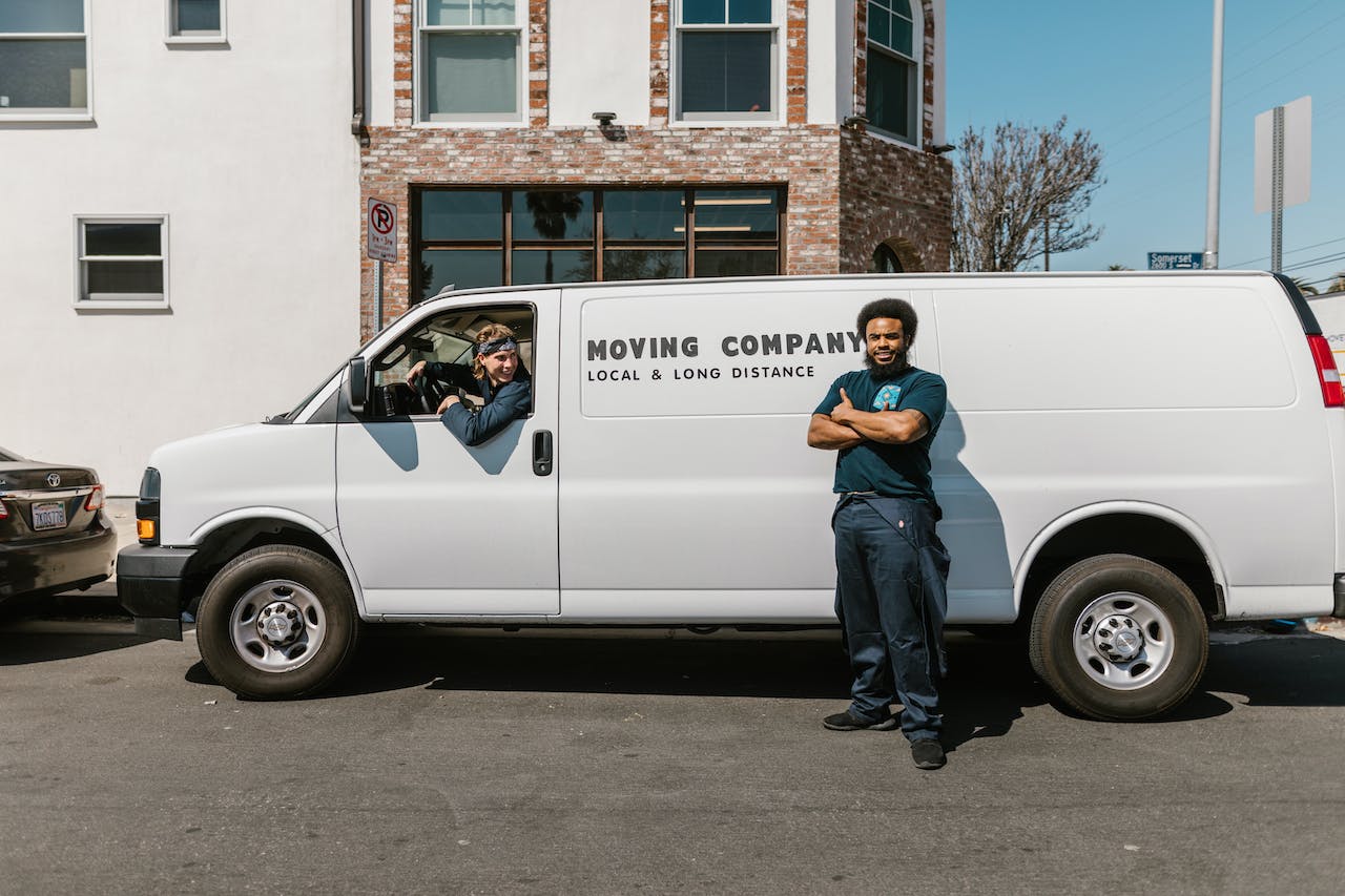 A moving company van with a man outside and a man inside the van posing for a picture.